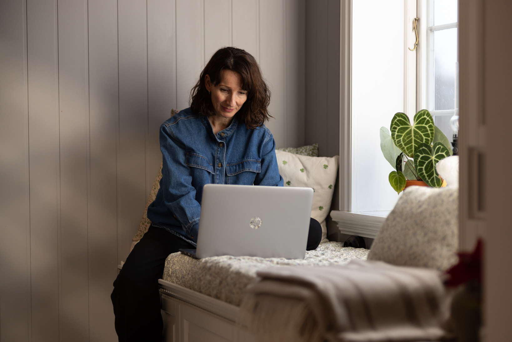 En kvinne som sitter på en benk inntil en vinduskarm med en bærbar PC